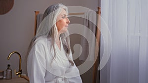 Pensive aged elderly lady 60s old Caucasian 50s senior mature gorgeous woman in white bathrobe standing in bathroom at