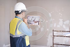 Pensive architect in white helmet looking at picture in hands stands against shabby wall