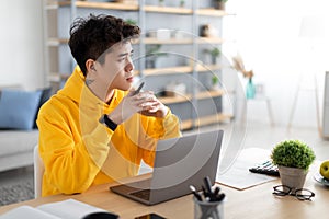 Pensive Asian man using laptop sitting at desk in office