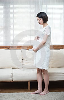 Pensive asian pregnant woman dreaming about child standing near window. Young happy expectant thinking about her baby and enjoying