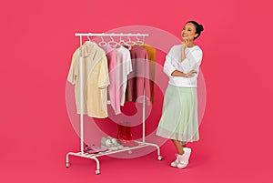 Pensive asian woman contemplating fashion choices, standing beside clothes rack