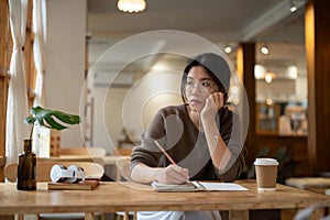 A pensive Asian woman sits at a table in a coffee shop, thinking while keeping her diary