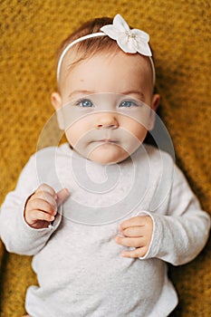 Pensive baby girl with a flower on her head in a white bodysuit lies on a yellow blanket, clenching her fists on her