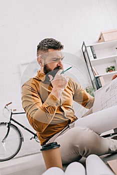 Pensive bearded adult male architect holding blueprint and working on project