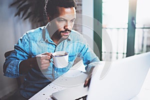 Pensive bearded African man using laptop home while drinking cup black coffee at the wooden table.Concept of young