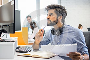 Pensive bearded business man with paper document and pen looking at computer screen and thinking of problem solution