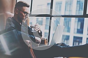 Pensive bearded businessman working at the modern loft office.Man sitting in vintage chair,holding in hands glass of