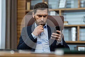 Pensive bearded guy in shirt and jacket holding cell phone and rubbing chin with worried facial expression. Concerned