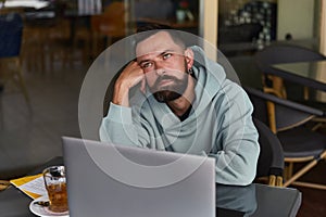 Pensive bearded man in casual clothes in cafe using his laptop