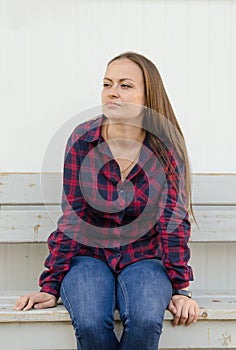Pensive beautiful girl with freckles with long hair and a red and blue checkered shirt and jeans sitting on bench