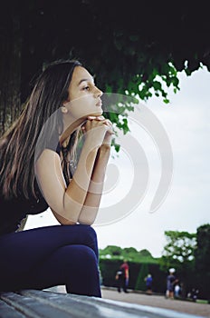 Pensive beautiful girl with long dark hair is sitting on a park bench