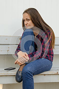 Pensive beautiful girl with long hair and a red and blue checkered shirt and jeans sitting on bench