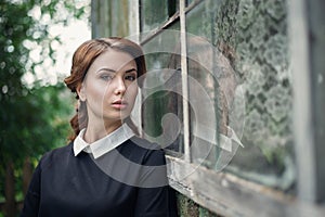 Pensive beautiful young girl in retro style dress standing near the window of old wooden house.