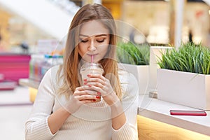 Pensive beautiful young woman drinking cocktail and thinking in cafe, looking down, hold cup with fresh baverage in hand, spend