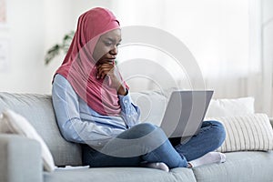 Pensive black muslim female freelancer with laptop at home brainstorming at project