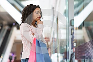 Pensive black shopaholic woman looking at showcase in department store, choosing clothes