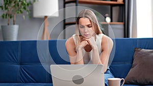 Pensive blonde woman thinking worried about problem sitting on couch in front of laptop pc at home