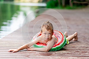 A pensive boy with a circle in the form of a watermelon in the summer lies on the shore of the lake, a child`s rest in the villag