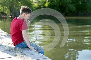 A pensive boy sits on a wooden pier, having lowered his feet in the water, and is nibbling a blade of grass.