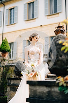 Pensive bride in a pink dress with a bouquet of flowers stands with her head bowed on the steps of an ancient villa