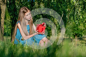 Pensive teen girl sitting on grass at city park and holding red gift box