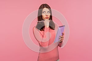 Pensive brunette woman in pink sweater holding notepad and pencil in hands thinking over plans for startup