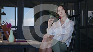 A pensive business woman sits on a chair at a table in the office and thinks dreamily.