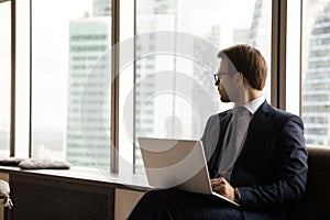 Pensive businessman with laptop sit in office looks skyscrapers city