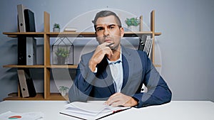 Pensive businessman with pen looking at camera near notebook.