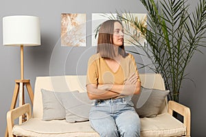 Pensive calm brown haired woman wearing beige T-shirt and jeans sitting on sofa in living room keeps hands folded looking away