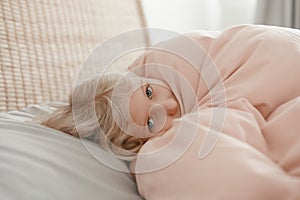 Pensive Caucasian blonde child girl lying on pillow under blanket in bed at home. Sad kid in stress or frustration. Child lying