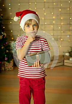 Pensive child in a red Santa Claus hat wondering what`s in the gift on the background of a Christmas tree. New Year`s