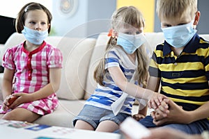 Pensive children are sitting on couch wearing protective medical masks and holding board playing cards