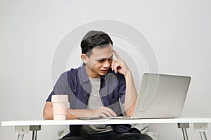 pensive confused asian man in the workplace whiile sitting in front of laptop computer. isolated background