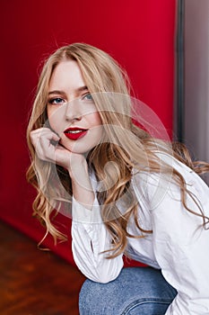 Pensive curly woman in shirt propping face with hand. Charming girl in jeans sitting on red background