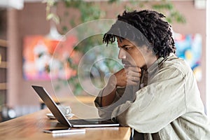Pensive curly young black man looks at laptop screen at table in cafe interior