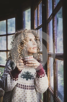 Pensive cute woman alone at home looking outside the window and drink cup of tea or coffee alone. Christmas holiday season. Young