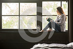 Pensive dreamy girl holding book sitting on sill at home photo