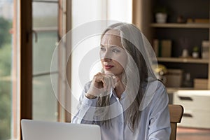 Pensive dreamy older 60s business lady sitting at laptop computer