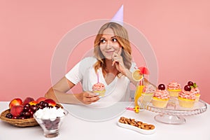 Pensive dreamy woman with blonde hair holding cake with candle and looking away with thoughtful expression, thinking about