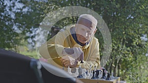 Pensive elderly man plays chess while sitting at table on nature background
