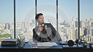 Pensive employee drinking coffee in panoramic office. Adult businessman resting