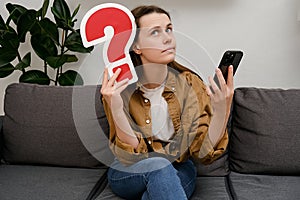 Pensive employee young caucasian woman 20s in shirt sitting on grey couch at home holding red question mark and smartphone. Online