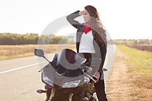 Pensive female driver in stylish clothes, poses on fast motorbike, looks thoughtfully aside, being active biker, poses in countrys