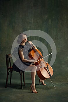 Pensive female musician, cellist sitting on chair with cello, in thoughtful pose against vintage green background