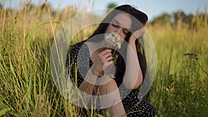 A pensive girl with dark long hair looks at white wildflowers, picks them and smells them enjoying the aroma while