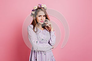 Pensive girl in flower wreath posing on pink background. Fascinating european lady in purple clothes standing in studio