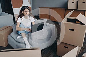 Pensive girl planning house interior design on laptop, sitting with boxes. Moving, flat renovation