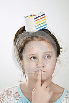 A pensive girl with a sguishy toy cake on her head and her index finger near the lips