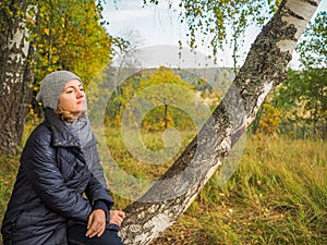 A pensive girl sitting on a birch tree on the outskirts of a withering autumn forest.